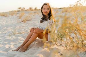 Summer photo of small girl in stylish boho outfit posing on the beach. Warm sunset colors. Wacation and  travel concept.