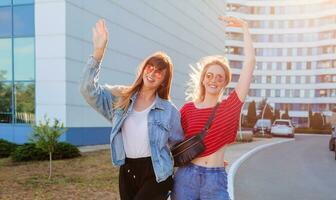 Two women, raised hands up and posing outdoor on urban background. Street style look. Sunset colors photo