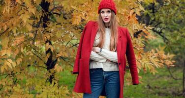 Autumn fashion portrait of blonde woman in red stylish coat and knitted hat walking in park. photo
