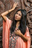 Beautiful asian model in pink boho dress posing over wood ornamental background. photo