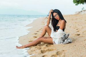 hermosa bronceado asiático mujer con rojo labios sentado en enviar cerca mar , en blanco playa bohemio atuendo . foto