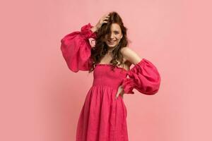 Stylish red-haired woman  playing with hairs and posing in pink lien dress with  sleeves over pink background. photo