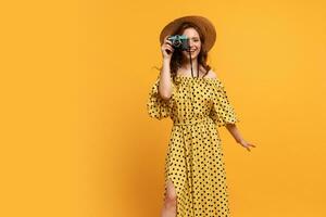 Travelling woman in straw hat and summer dress posing with retro camera over yellow backgrond in studio. photo