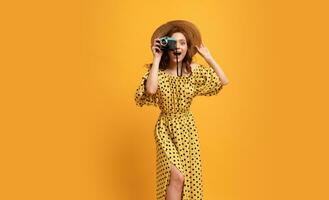 Lovely red head woman in straw hat and stylish summer dress posing over yellow background isolate in studio. Holding retro camera. photo