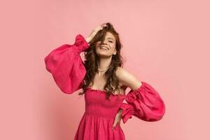 Stylish red-haired woman  playing with hairs and posing in pink lien dress with  sleeves over pink background. photo