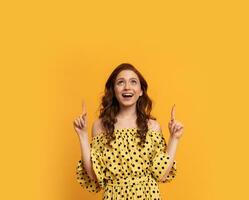 Red  head woman with suprice face pointing up   in yellow  summer dress posing over yellow background in studio. Summer style. photo
