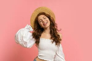 juguetón mujer en elegante lino parte superior con globo mangas posando en rosado antecedentes. ondulado pelos, Paja sombrero, verano Moda tendencias foto