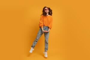 Laughing stylish red head  woman in orange sweater and blue jeans  jumping  over  yellow background.  Full lenght. photo