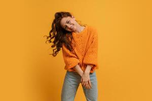 Excited young girl in  orange  sweater  fooling around in studio jumping with fluttering hair isolated on yellow rbackground. People sincere emotions lifestyle. photo