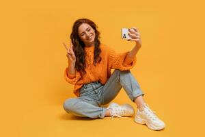 Attractive red head woman in stylish fall outfitm sweater and jeans posing over  yellow  background in studio.   Making self portrait by  smartphone. photo