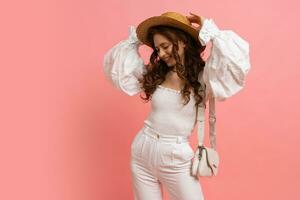 Lovely woman in elegant white  linen top with balloon sleeves posing on pink background.  Wavy hairs, straw hat, summer fashion trends. photo