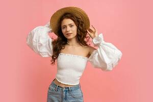 Happy Woman in summer outfit plays hair on pink background. Straw hat, white top with sleeves, natural make up. photo