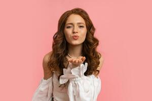 Smiling woman with perfect wavy hairstyle in white top wiyh sleeves  send kiss to camera on pink background. Isolate. photo