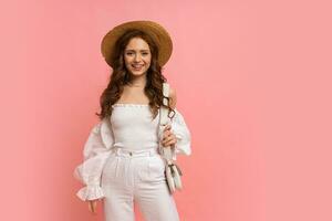 encantador mujer en elegante blanco lino parte superior con globo mangas posando en rosado antecedentes. ondulado pelos, Paja sombrero, verano Moda tendencias foto