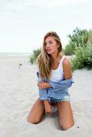 Close up portrait of blond seductive  girl with long shiny wavy hairs on summer beach enjoying vacation.  Striped blouse, jeans shorts. Straw hat. photo