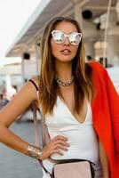 Blond woman in big sunglasses  with full lips posing outdoor. Red jacket, stylish silver accessorises. photo