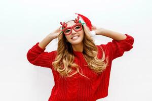 alegre despreocupado rubio mujer en linda mascarada lentes y nuevo año sombrero en rojo de punto suéter posando en blanco antecedentes . aislar. Navidad y nuevo prueba fiesta concepto. foto