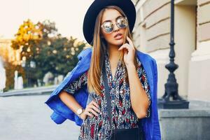 cerca arriba soleado estilo de vida retrato de elegante casual mujer en negro sombrero, brillante vestir y azul chaqueta en espalda caminando en europeo calles Moda y compras concepto. foto