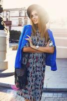 Close up sunny  lifestyle portrait of elegant casual woman in black hat, bright dress and blue jacket  on shoulders  walking   on european streets. Fashion and shopping   concept. photo