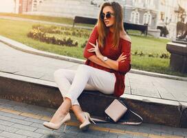 bonito elegante mujer en de moda Gafas de sol y rojo suéter sentado y flirteador . al aire libre calle estilo retrato. alegre mujer vistiendo tacones y blanco vaqueros. foto