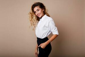 Lovely blond woman with perfect smile in white blouse posing over beige background in studio. Stylish workwear outfit. photo