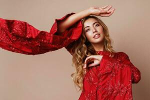 Seductive blonde woman posing in luxury red dress with wide sleeves in studio. Fashionable look. Blond wavy hairs. Expressive photo. Windy cloth. photo