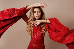 Studio fashion portrait of young blond lovely woman in luxurious red dress with wide sleeves.  Expressive pose. Beige background. photo