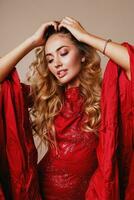 Close up beauty portrait of perfect blond woman with full lips, natural make up posing in studio in amazing luxury red dress with sequins and wide sleeves. Beige background. Hands near face. photo