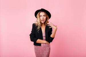 Girl with surprise face standing over pink background . Wearing elegant dress with sequins . Amazed emotions.Wearing trendy dress with sequence , black jacket and hat. photo