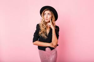 Pensive blonde woman in black hat posing over pink background. Wearing trendy dress with sequence , black jacket and hat. photo