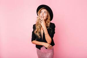 Pensive blonde woman in black hat posing over pink background. Wearing trendy dress with sequence , black jacket and hat. photo