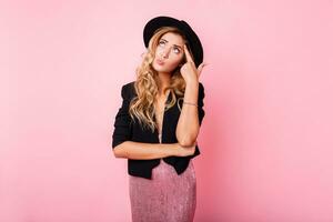 Pensive blonde woman in black hat posing over pink background. Wearing trendy dress with sequence , black jacket and hat. photo