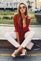 Pretty stylish  woman in trendy sunglasses and red sweater sitting and   flirting . Outdoor street style portrait. Cheerful woman wearing  heels and white jeans. photo