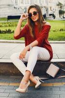 Pretty stylish  woman in trendy sunglasses and red sweater sitting and   flirting . Outdoor street style portrait. Cheerful woman wearing  heels and white jeans. photo