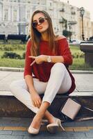 Pretty stylish  woman in trendy sunglasses and red sweater sitting and   flirting . Outdoor street style portrait. Cheerful woman wearing  heels and white jeans. photo