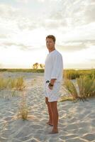 Attractive handsome european man standing on the beach. Sunset. White clothes. Summer fashion. photo