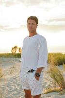 Attractive handsome european man standing on the beach. Sunset. White clothes. Summer fashion. photo