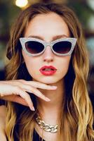Close up fashion portrait of glamour stylish  blonde  lady   with cool retro sunglasses . The girl sitting in cafe and enjoying  time out. photo