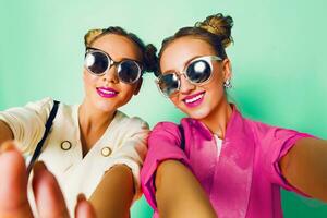 Fashion studio image of two young women in stylish casual  spring outfit   having fun, show  tongue. Bright trendy pastel  colors, stylish hairstyle  with buns , cool sunglasses. Friends portrait. photo