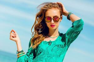 al aire libre Moda verano retrato de hermosa Delgado bronceado mujer con Perfecto presagiar en brillante boho playa vestir y frio accesorios , elegante Gafas de sol en el paraíso tropical incumplimiento. foto