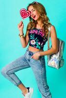 Studio   positive portrait of young sexy funny fashion crazy woman posing on blue wall background in summer style outfit with pink lollipop wearing print  top , neon   backpack  and cute glasses. photo