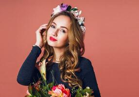 beautiful young female with long wavy blonde  hair in wreath of spring flowers  posing with flower bouquet over   pink  background. photo