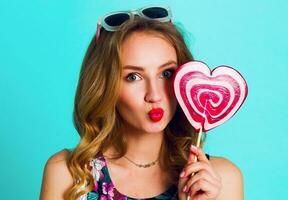 Studio   positive portrait of young sexy funny fashion crazy woman posing on blue wall background in summer style outfit with pink lollipop wearing print  top , neon   backpack  and cute glasses. photo