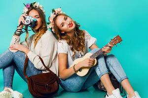 two young funny fashion girls  posing on blue wall background in summer style outfit with  flowers  wreath wearing blue jeans and boho bag pack. . photo