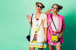 Fashion studio image of two young women in stylish casual  spring outfit   having fun, show  tongue. Bright trendy   colors, stylish hairstyle  with buns , cool sunglasses. Friends portrait. photo