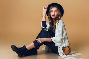 High fashion portrait of young elegant blonde  woman in black wool hat  wearing oversize white fringe  poncho with long grey dress. Studio shot. American hippie bohemian style. Beige background. photo