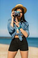 sexy mujer con Perfecto bronceado cuerpo, lleno rojo labios y l largo piernas posando en el tropical soleado playa. vistiendo cosecha parte superior , pantalones cortos y Paja sombrero. brillante soleado colores. foto