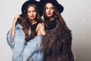 Positive fashion portrait of two girls, best friends posing indoor on bright  greu background wearing winter stylish fluffy coat, black casual hat. Fashionable clothes.  Sisters walking. photo