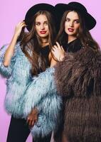 positivo Moda retrato de dos chicas, mejor amigos posando interior en brillante rosado antecedentes vistiendo invierno elegante mullido abrigo, negro casual sombrero. de moda ropa. hermanas caminando. foto