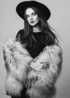 Close up indoor studio fashion portrait of pretty young model in stylish winter fluffy coat and black hat posing . Evening bright make up. Black and white. Monochrome colors. photo